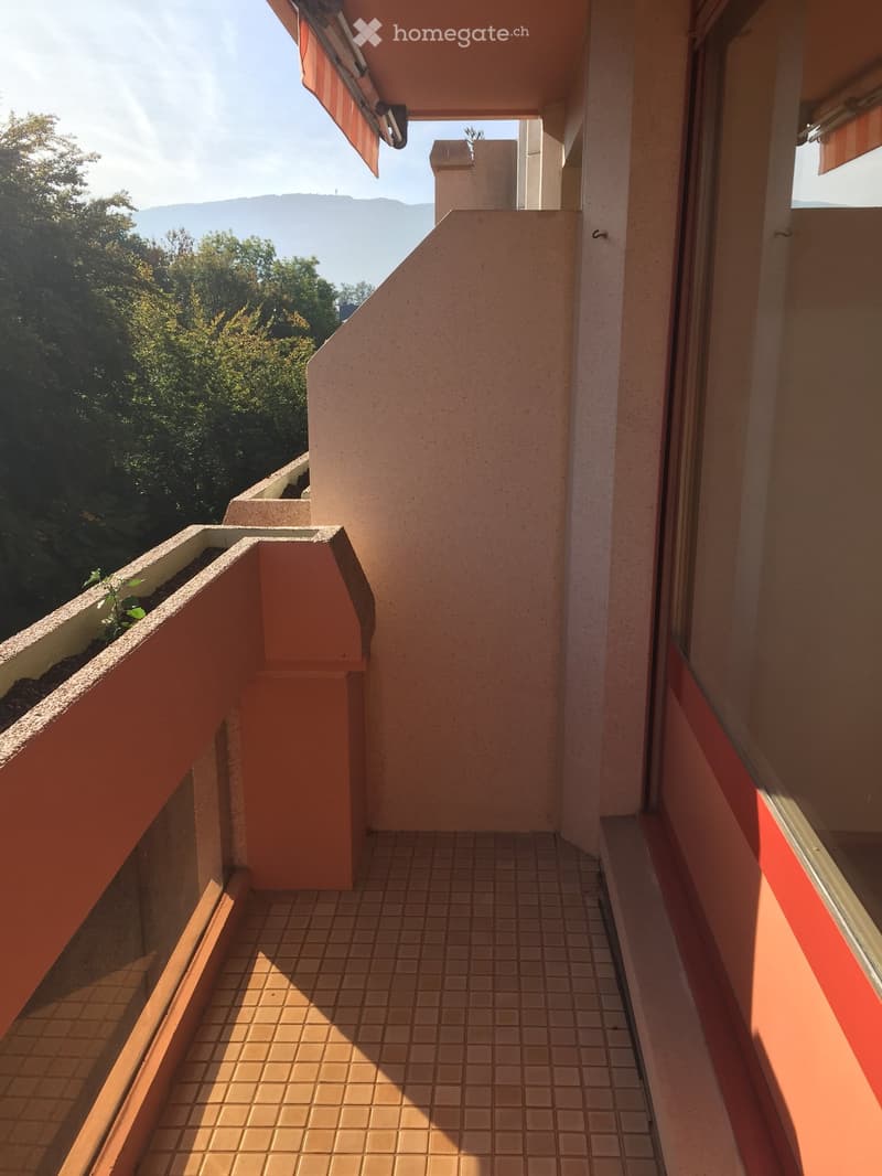 Balcony with tiled floor, beige walls, and a view of greenery and mountains in the background.