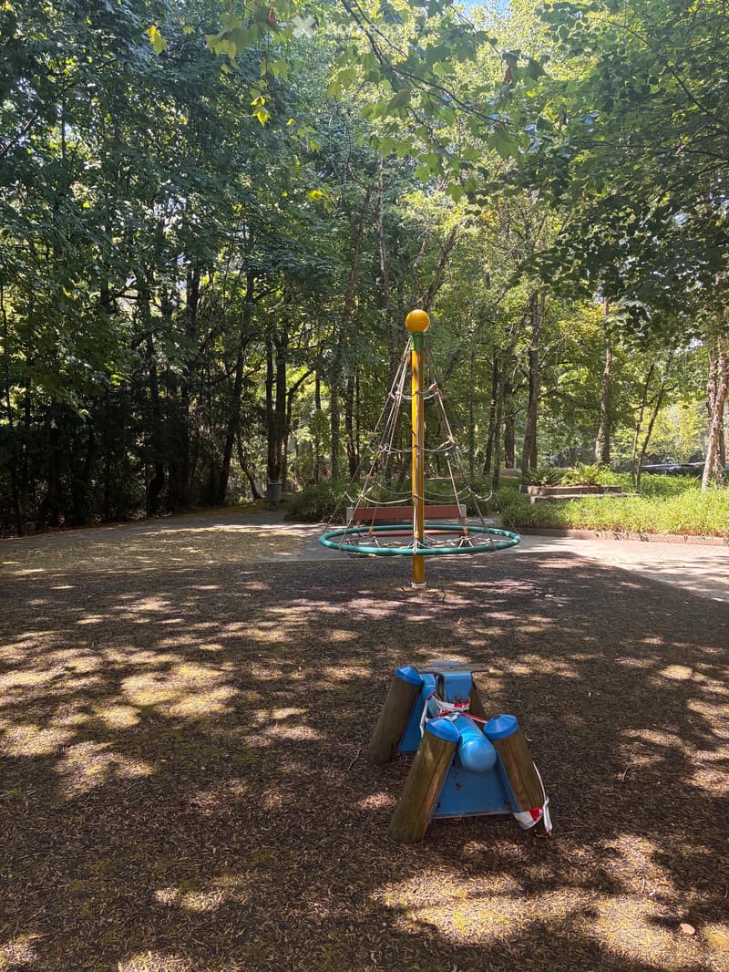 A playground area with a round nest swing and a blue wooden seesaw, surrounded by trees and a gravel path.