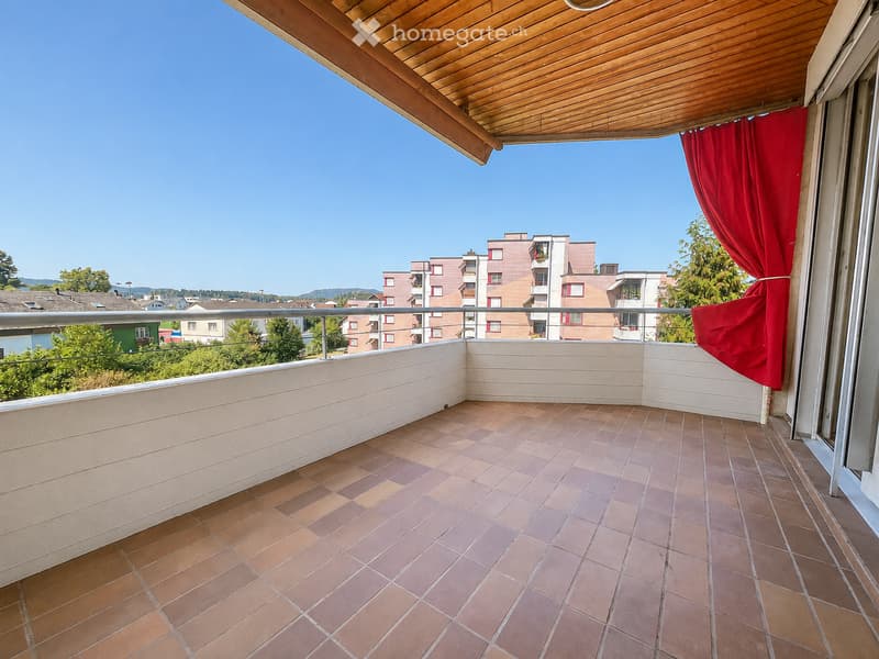 Balcony with tiled floor, wooden ceiling, metal railing, and red curtain on one side, overlooking a residential area with buildings and trees.