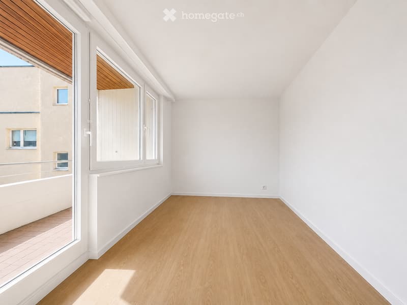 Empty room with light wooden flooring, white walls, and large windows with wooden frames, leading to a balcony area.