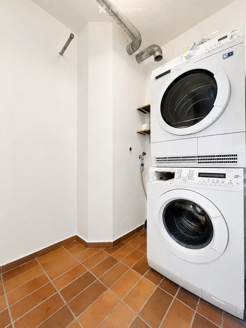A small, well-lit room with white walls and orange tiled flooring, featuring a stacked washing machine and dryer unit.