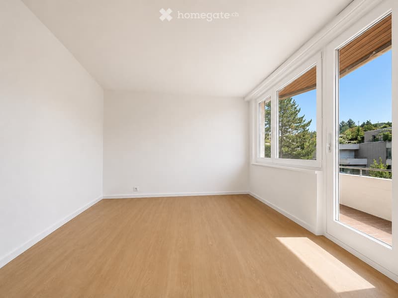 Empty room with light wooden flooring, white walls, large windows, and a sliding glass door leading to a balcony.