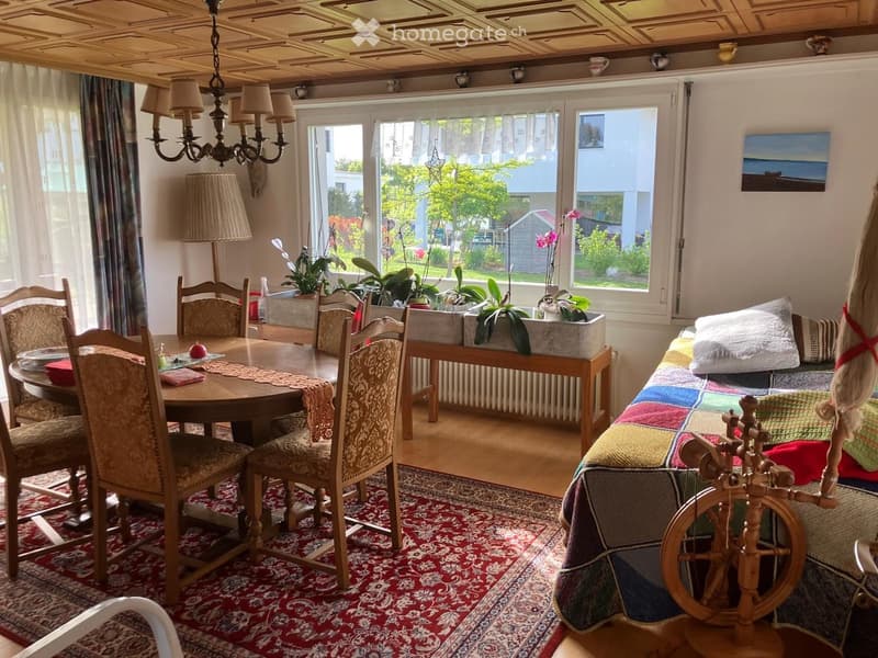 Bright living and dining area with wooden furniture, a patterned rug, a chandelier, and a spinning wheel near the sofa.