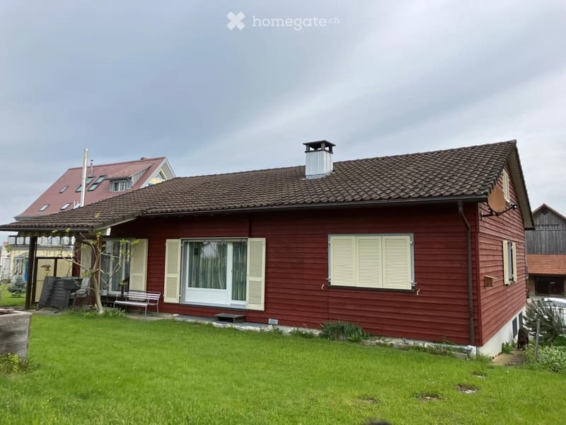 Red wooden house with a tiled roof, white shutters, and a chimney. The house has multiple windows and a small porch area with outdoor furniture.
