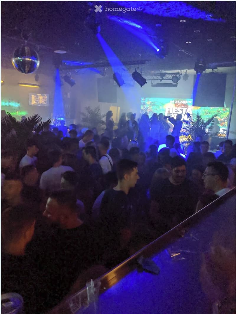Crowded nightclub with blue and red lighting, disco ball, bar area, large screen displaying the word Fiesta