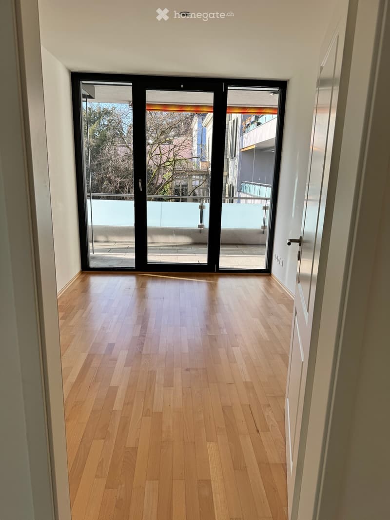 Empty hallway with light wooden flooring, white walls, and large sliding glass doors leading to a balcony area.