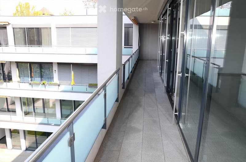 Modern hallway with tiled flooring, glass railings, and multiple levels visible with balconies and windows.