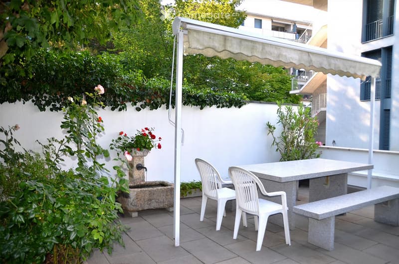 A small outdoor patio with a table, two white chairs, a bench, and a fountain, surrounded by greenery and a white wall.