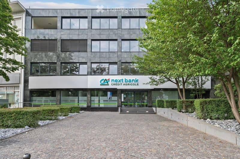 Modern multi-story building with large windows and a sign reading "next bank CREDIT AGRICOLE" above the entrance. The building has a sleek, contemporary design with a stone facade and is surrounded by greenery and a paved entrance area.