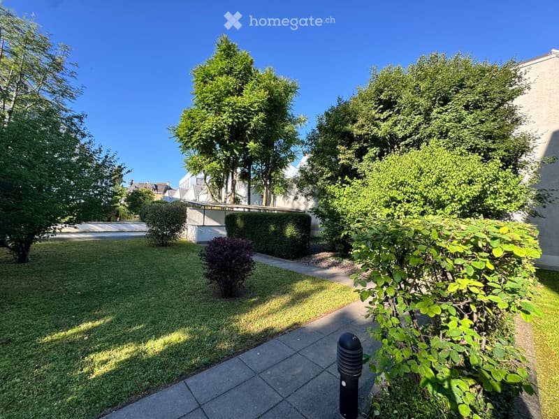 Well-maintained garden with neatly trimmed hedges, trees, and a paved pathway leading to a building entrance.