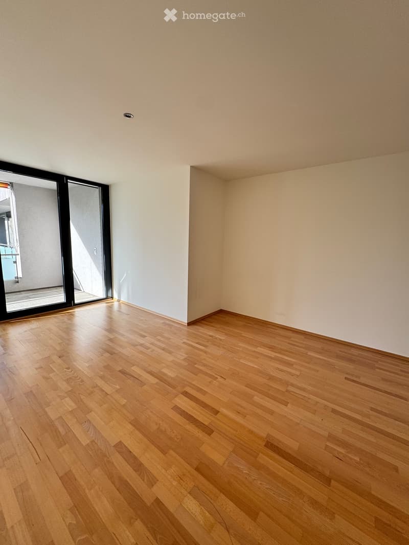 Empty room with light wooden flooring and white walls, featuring a large sliding glass door leading to a balcony.