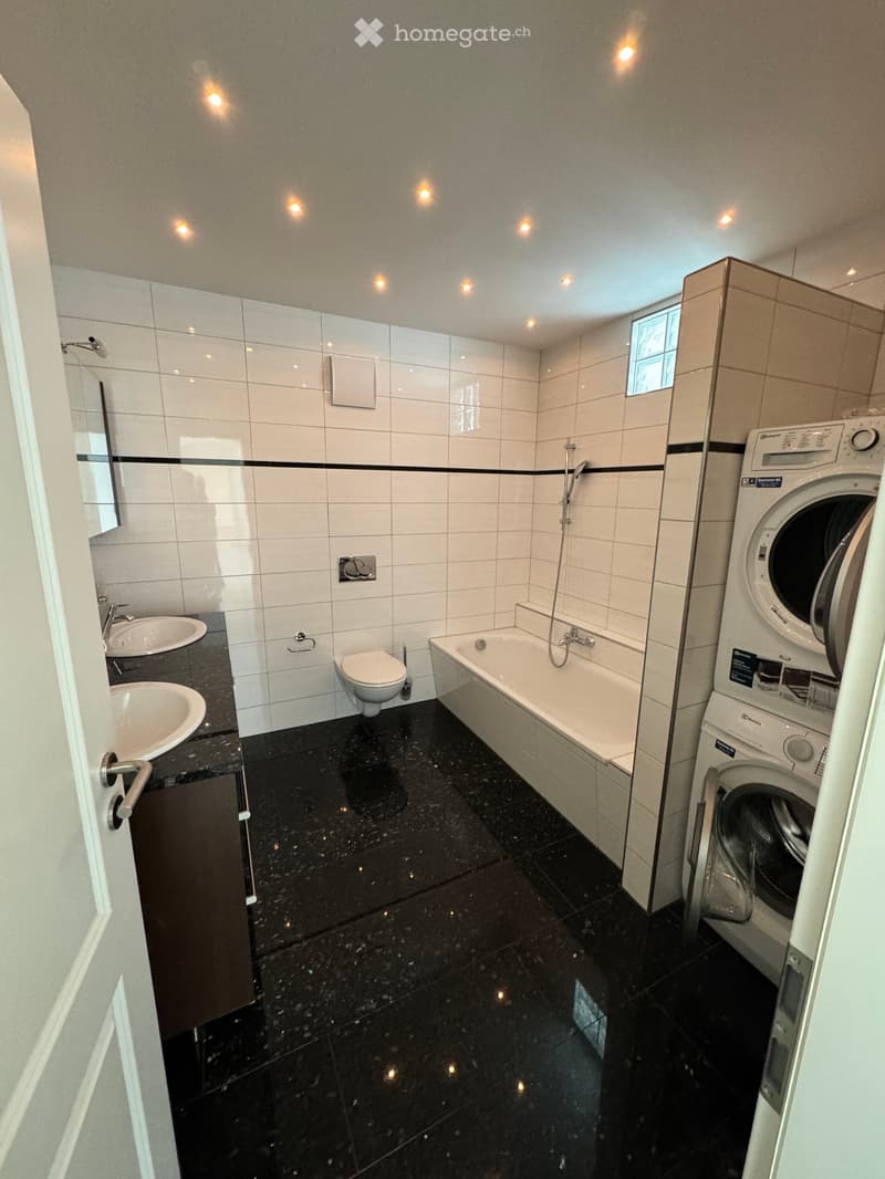 Modern bathroom with black marble flooring, white tiled walls, a bathtub, toilet, sink, and a washing machine.