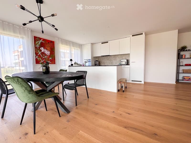 Open-plan living and dining area with a round wooden dining table, green upholstered chairs, modern kitchen with white cabinets, and a sleek chandelier.
