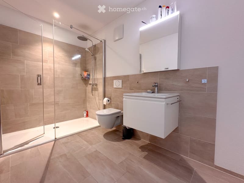 Modern bathroom with beige tiles, a glass-enclosed shower, a white toilet, a sink with a mirror, and a small shelf with toiletries.