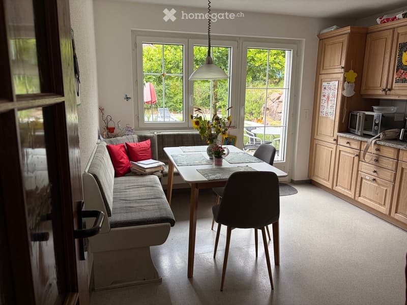 Bright living and dining area with a grey sofa, red cushions, a dining table with chairs, and a kitchen area with wooden cabinets and a microwave.