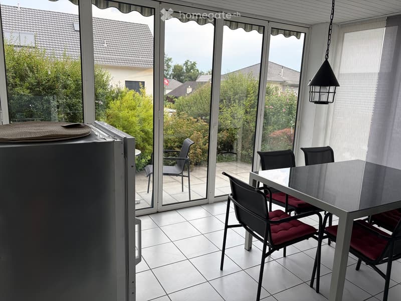 Dining area with a white table, red cushioned chairs, and a refrigerator, connected to a patio with outdoor seating.