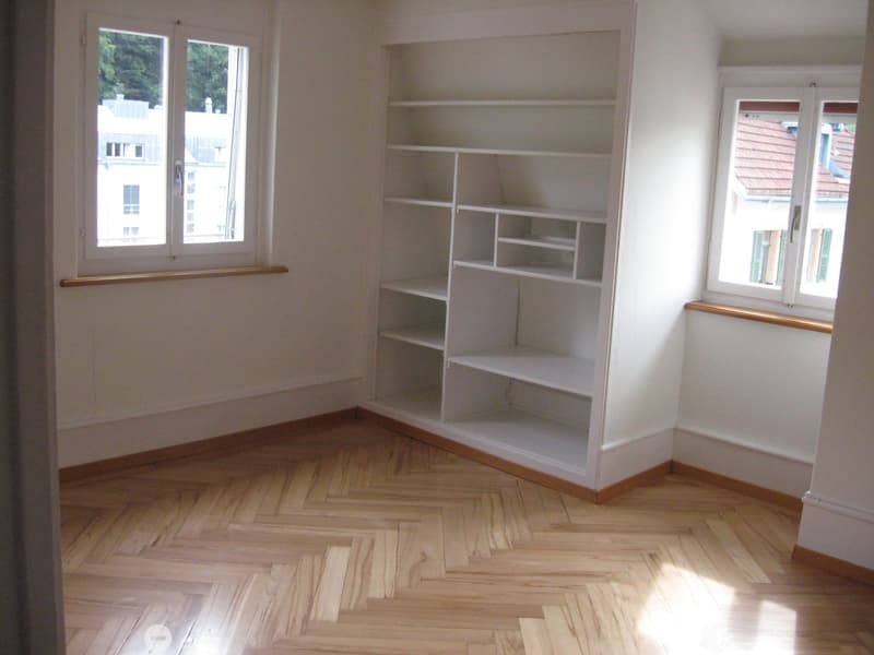 Empty room with wooden herringbone flooring, white walls, built-in shelving unit, and large windows with wooden sills.