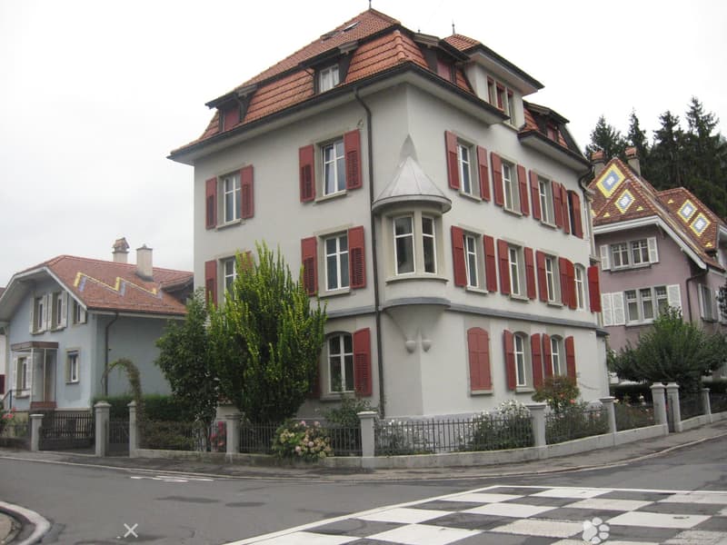 A three-story residential building with a red-tiled roof, white walls, and red shutters. The building features a bay window and is surrounded by a low fence with decorative plants and flowers.