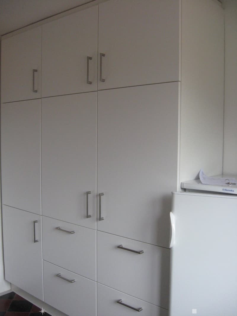 Modern kitchen with white cabinets and a stainless steel refrigerator. The cabinets have sleek, minimalist handles and are arranged in a vertical stack.