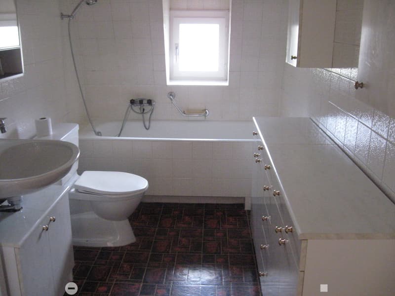 Bathroom with white tiles, a toilet, sink, bathtub, and a window above the bathtub. The floor has dark tiles.