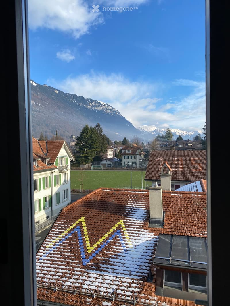 View from a balcony showing a town with red-tiled roofs, green spaces, and snow-capped mountains in the background.