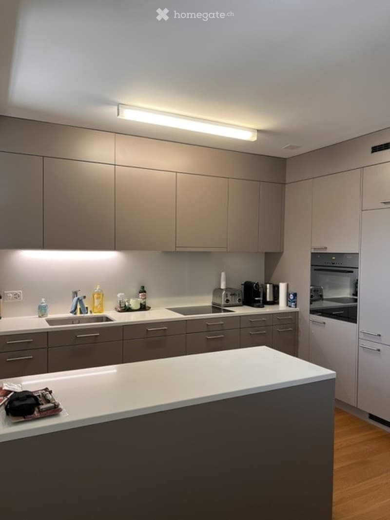 Modern kitchen with grey cabinets, white countertops, and stainless steel appliances including a sink, fridge, and oven. A coffee maker and various bottles are placed on the counter.