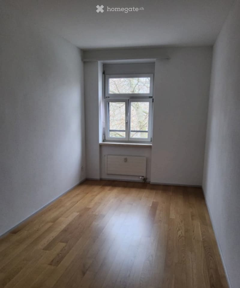 Empty room with wooden flooring, white walls, and a single window with a radiator below it.