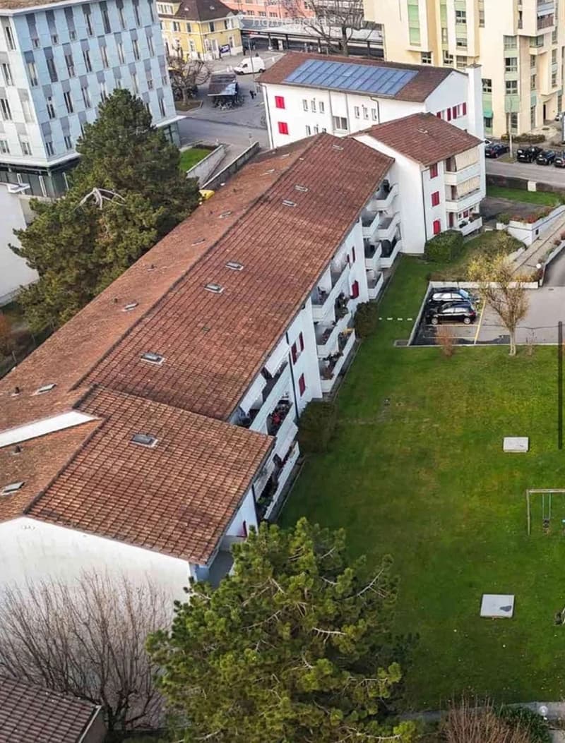 Aerial view of a multi-story residential building with red-tiled roofs, balconies, and surrounding greenery. A parking area is visible in the foreground.