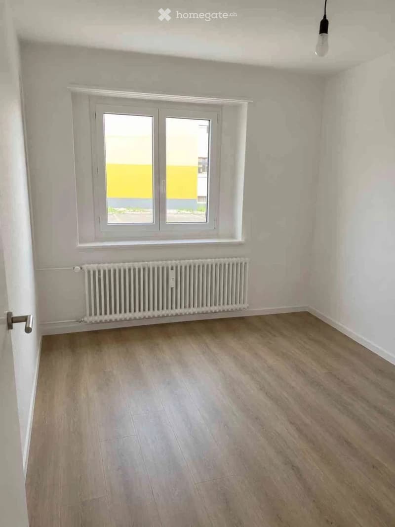 Empty room with light wooden flooring, white walls, a window with white frame, radiator, and a door with a silver handle.