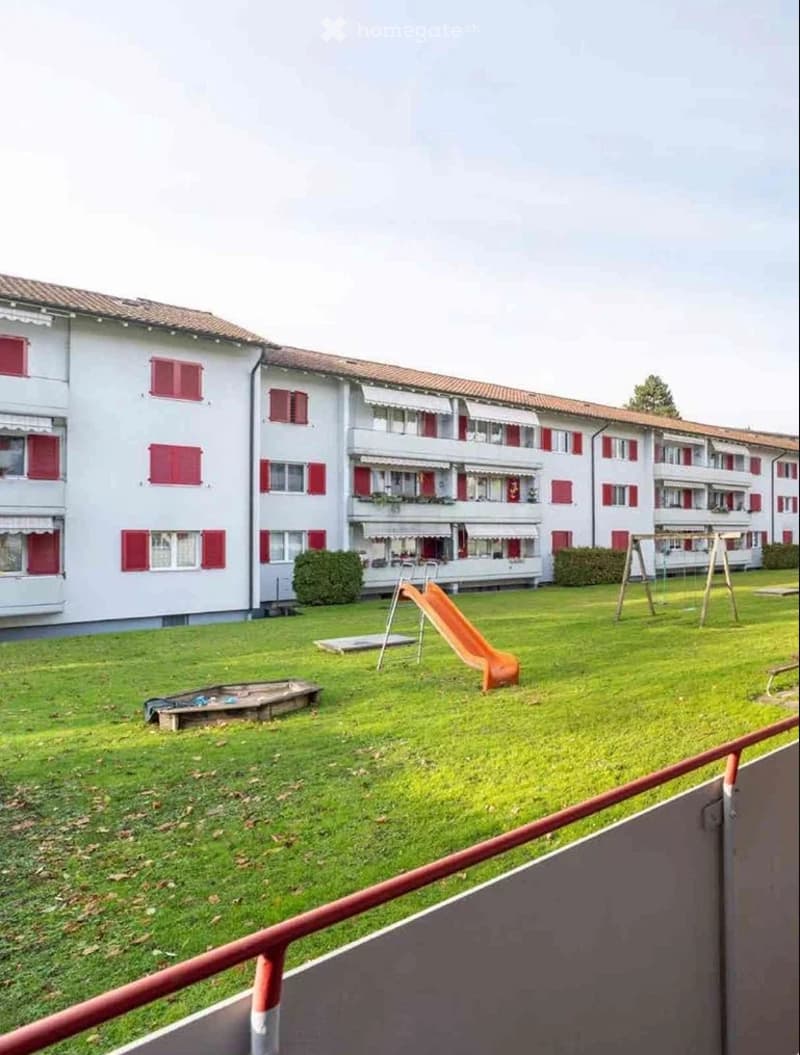 A grassy outdoor area in front of a multi-story residential building with red shutters. Features include a playground with a slide, a sandbox, and a swing set.
