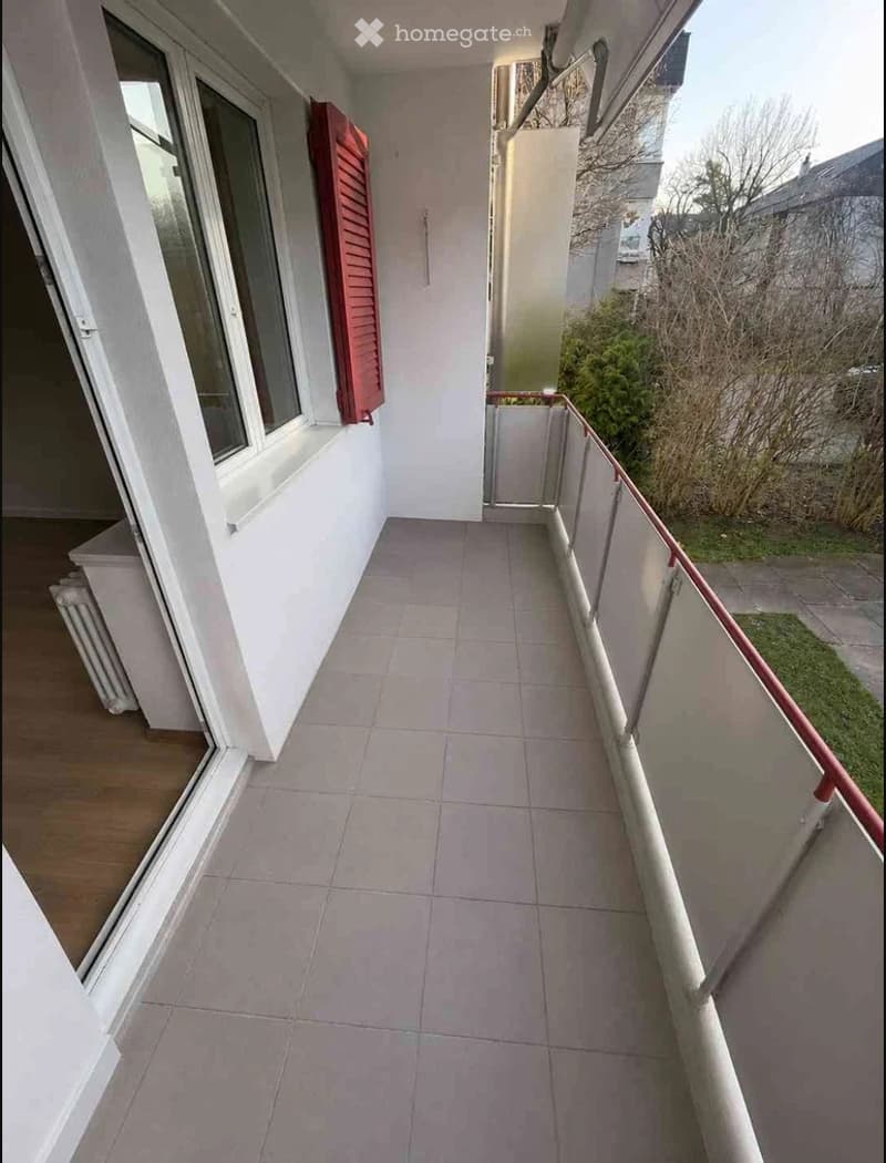 Balcony with tiled floor, white walls, red shutters, and a metal railing. The balcony overlooks a small garden area with bushes and grass.