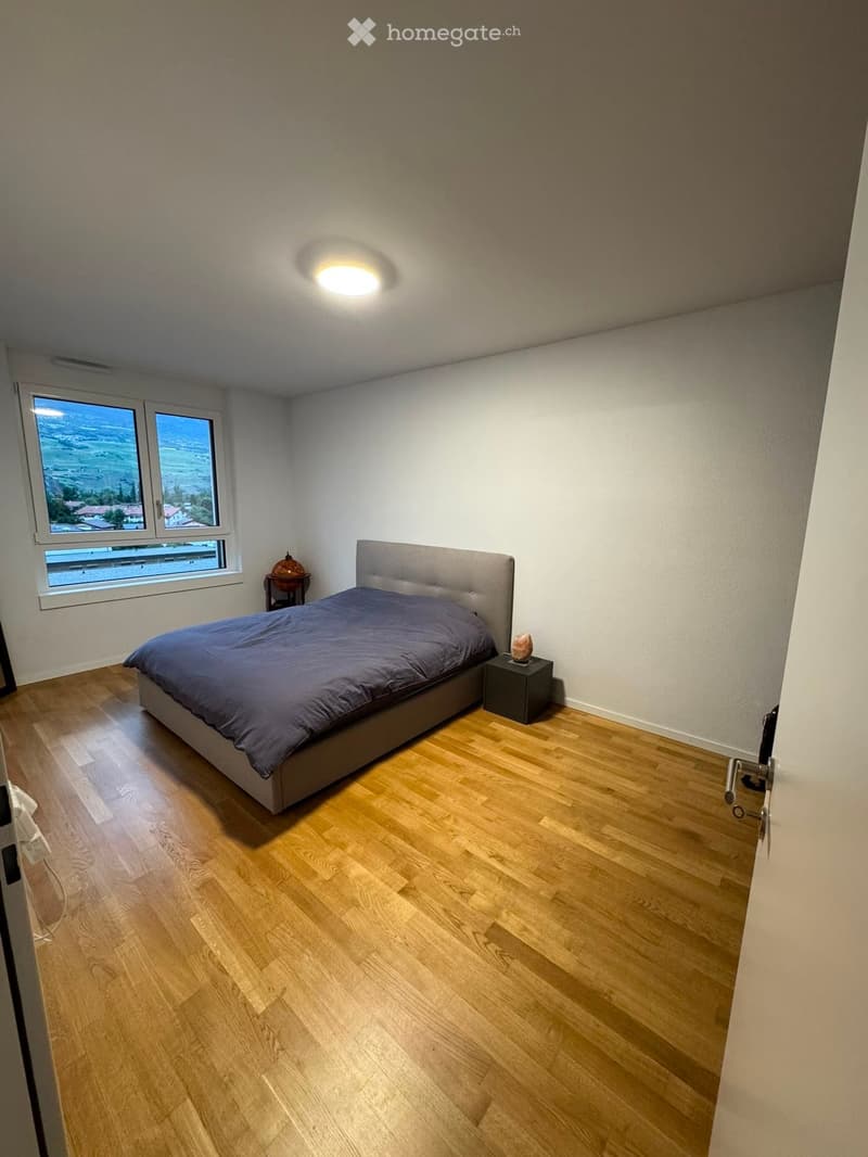 Modern bedroom with wooden flooring, large window offering a scenic view, a neatly made bed with a dark blue duvet, a nightstand with a table lamp, and a decorative globe on a small table.