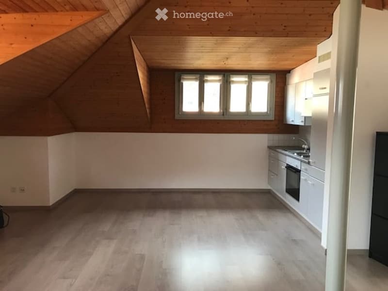 Empty attic space with wooden ceiling beams, large window, and white kitchen cabinets.