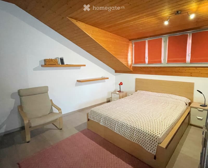 Cozy attic bedroom with wooden slanted ceiling, bed, armchair, nightstand, and built-in shelves.