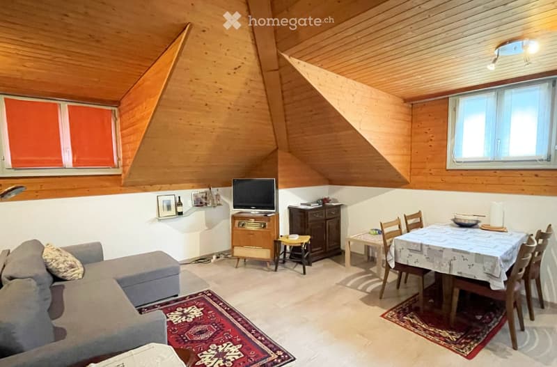 Cozy attic room with wooden slanted ceilings, large windows, a sofa, a dining table with chairs, a TV, and a small cabinet.