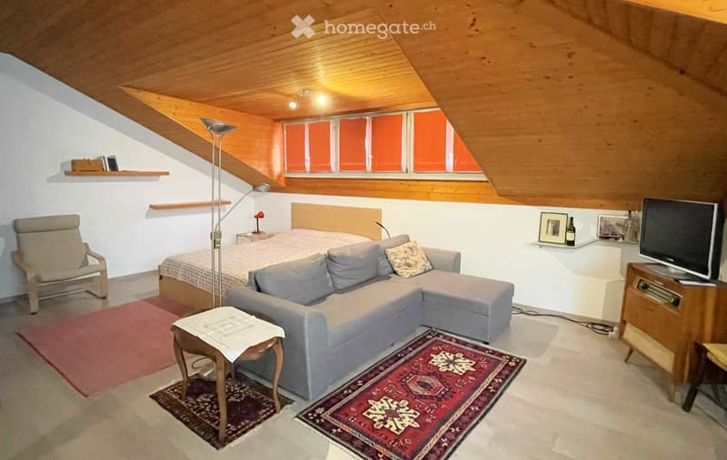 Cozy attic room with wooden slanted ceilings, white walls, and large windows. Features a grey sectional sofa, a wooden armchair, a small table with a white cloth, a television on a wooden stand, and a bed with a wooden headboard.