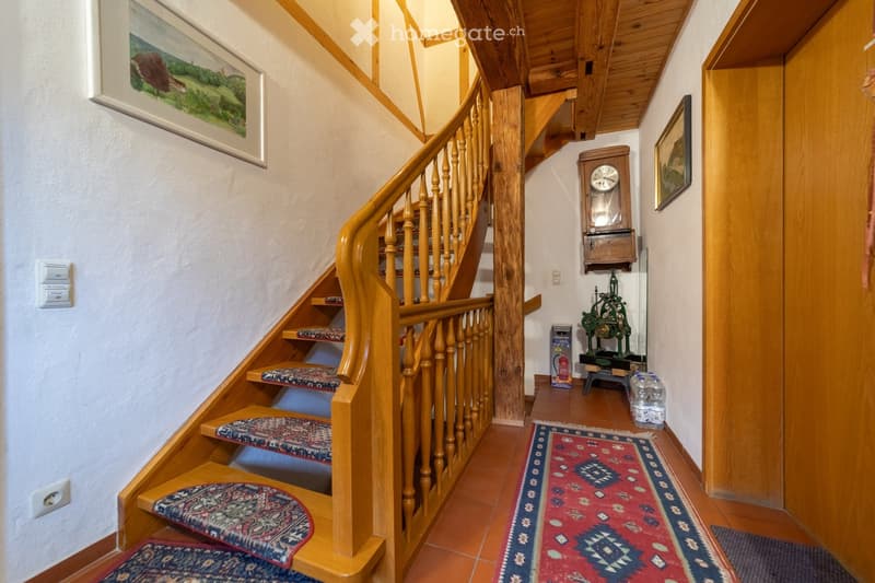 Wooden staircase with patterned carpeted steps, wooden handrail, and a wooden clock on the wall. The hallway features a red and blue patterned rug, a small green machine, and framed artwork on the walls.