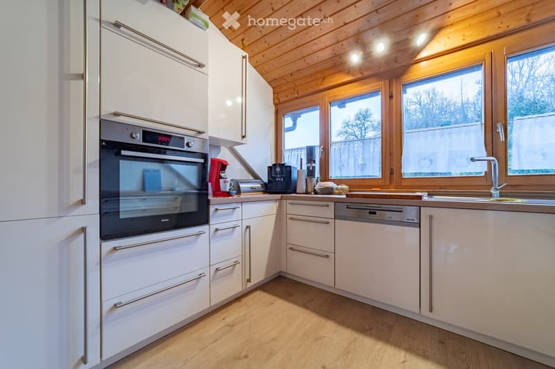 Modern kitchen with white cabinets, wooden ceiling, and large windows. Features an oven, dishwasher, coffee maker, and sink.
