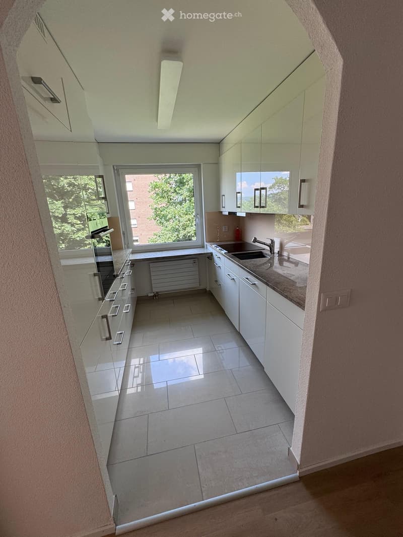 Modern kitchen with white cabinets, tiled floor, and a large window overlooking greenery. Features a sink, faucet, and radiator.