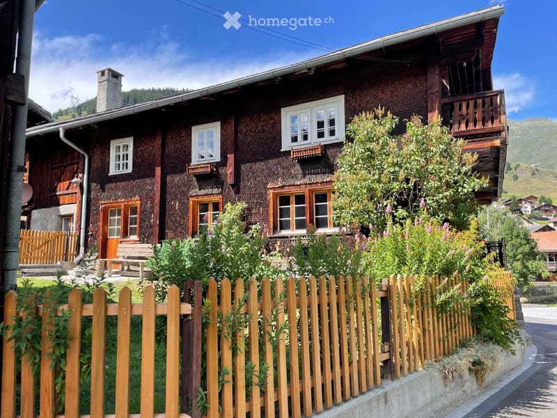 Traditional wooden house with multiple windows, a balcony, and a wooden fence in front, surrounded by greenery and a scenic mountain backdrop.