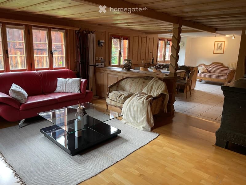 Cozy living room with wooden beams, large windows, red leather sofa, glass coffee table, and a patterned armchair with a blanket.
