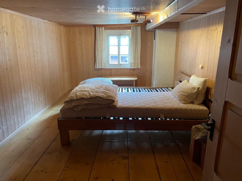 Cozy wooden bedroom with a single bed, pillows, blankets, a small bench, and a table near the window.
