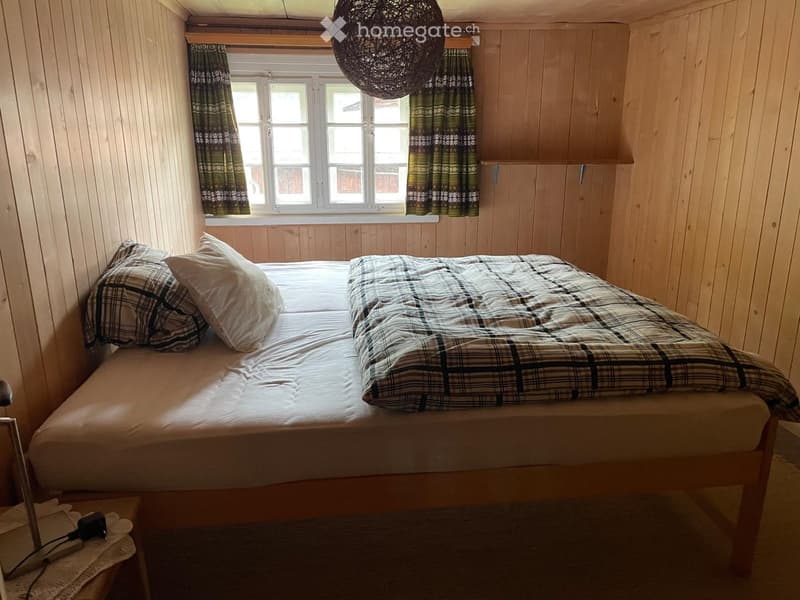Cozy wooden bedroom with a single bed, plaid blanket, pillows, and a window with curtains. A small table with a lamp is visible on the left side.
