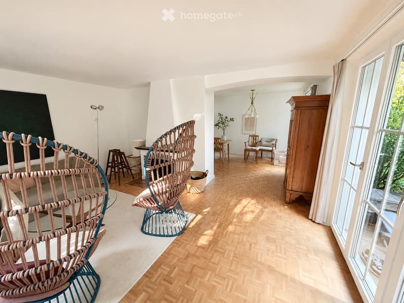 Modern living room with wooden herringbone flooring, large windows, and contemporary furniture including a woven sofa, chairs, a coffee table, and a wooden cabinet.