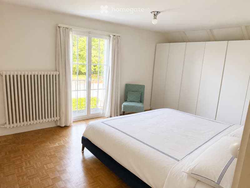 Elegant bedroom with a large bed, a matching chair, a tall wardrobe, and a radiator. The room features wooden flooring, large windows with curtains, and a modern ceiling light.
