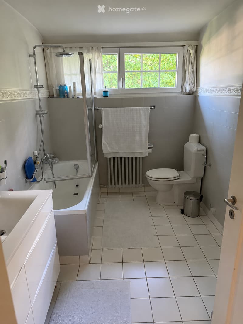 Bathroom with white tiled walls and floor, bathtub with shower, toilet, sink, towel, radiator, and trash can.