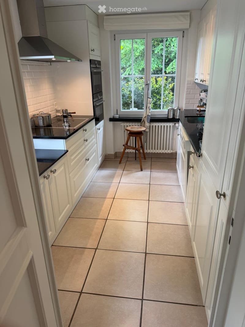Modern kitchen with white cabinets, black countertops, tiled floor, and a window overlooking greenery. Includes an oven, sink, and a wooden stool.
