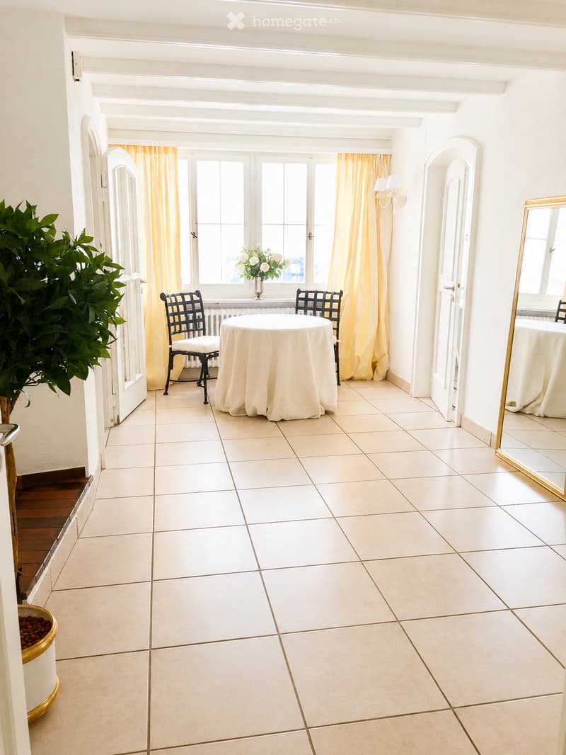 Bright, tiled room with a round table, two chairs, a large mirror, a vase with flowers, and a potted plant near the doorway.