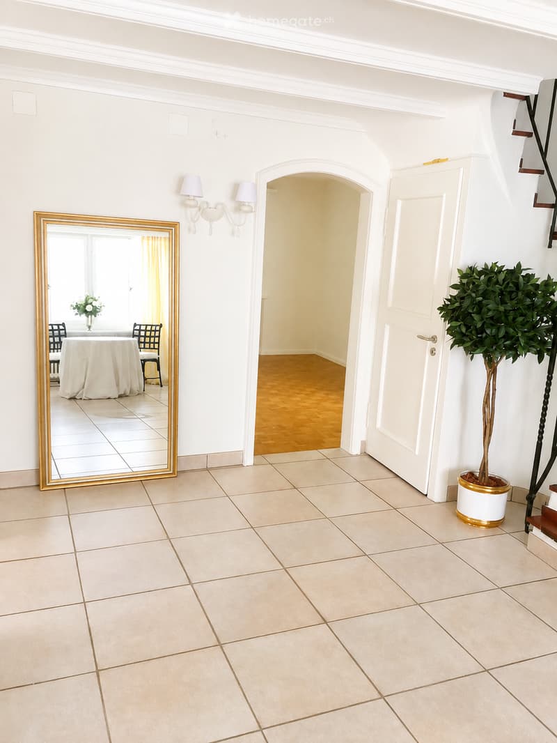 Empty hallway with tiled floor, white walls, arched doorway leading to another room, large mirror, and potted plant.