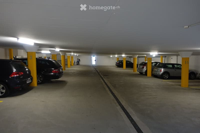 Underground parking area with several parked cars, yellow support pillars, and numbered parking spots. The area is well-lit with overhead lighting and has a concrete floor.